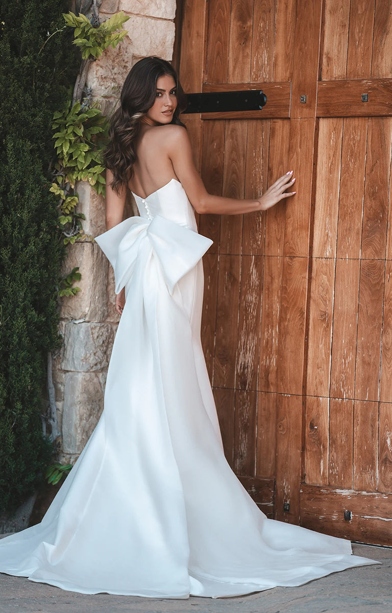Woman in A1216 by Allure Bridals - Strapless Stretch Mikado Gown with Oversized Bow with a large bow standing in front of a wooden door.