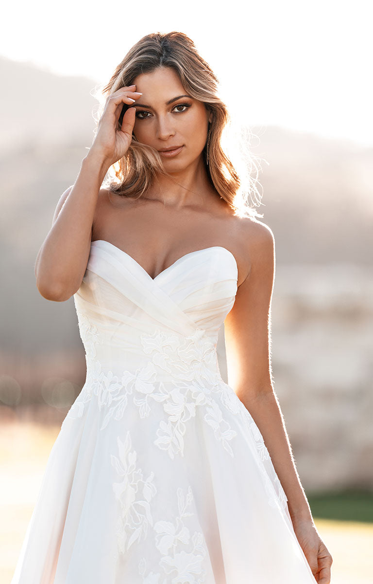 Woman wearing A1257 by Allure Bridals - Strapless Sweetheart Tulle Ballgown with Floral Lace Appliqués standing on grass with a rustic whitewashed brick wall behind.