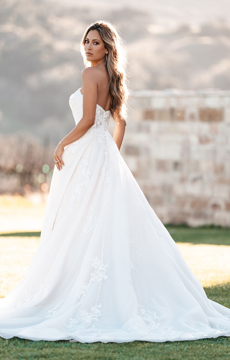 Woman wearing A1257 by Allure Bridals - Strapless Sweetheart Tulle Ballgown with Floral Lace Appliqués standing on grass with a rustic whitewashed brick wall behind.