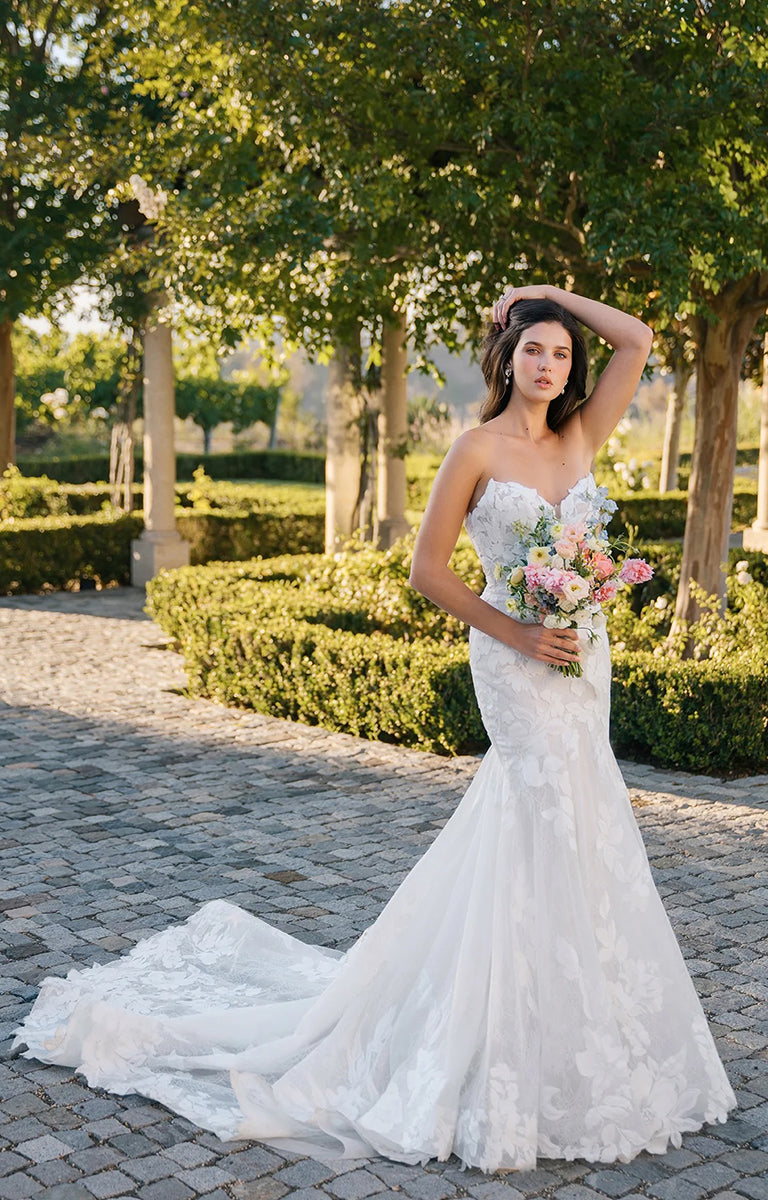 Woman in A1401 by Allure Bridals - Matte Lace Fit-and-Flare Wedding Dress with Cat-Eye Neckline holding a bouquet of flowers in a garden setting