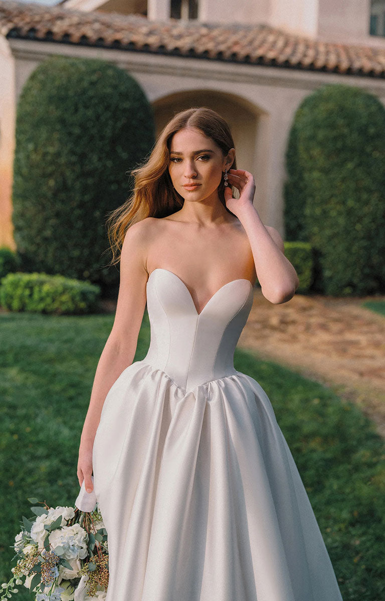 A woman wearing A1492 by Allure Bridals - Mikado Ball Gown with Basque Waist, Sweetheart Neckline and Detachable Panniers, structured bodice, and full skirt, standing outdoors in front of a villa with manicured hedges and holding a bouquet of white flowers.