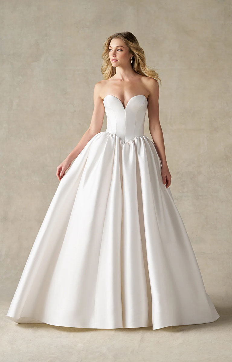 A woman modeling A1492 by Allure Bridals - Mikado Ball Gown with Basque Waist, Sweetheart Neckline and Detachable Panniers wedding dress in white satin, standing against a textured beige studio backdrop.