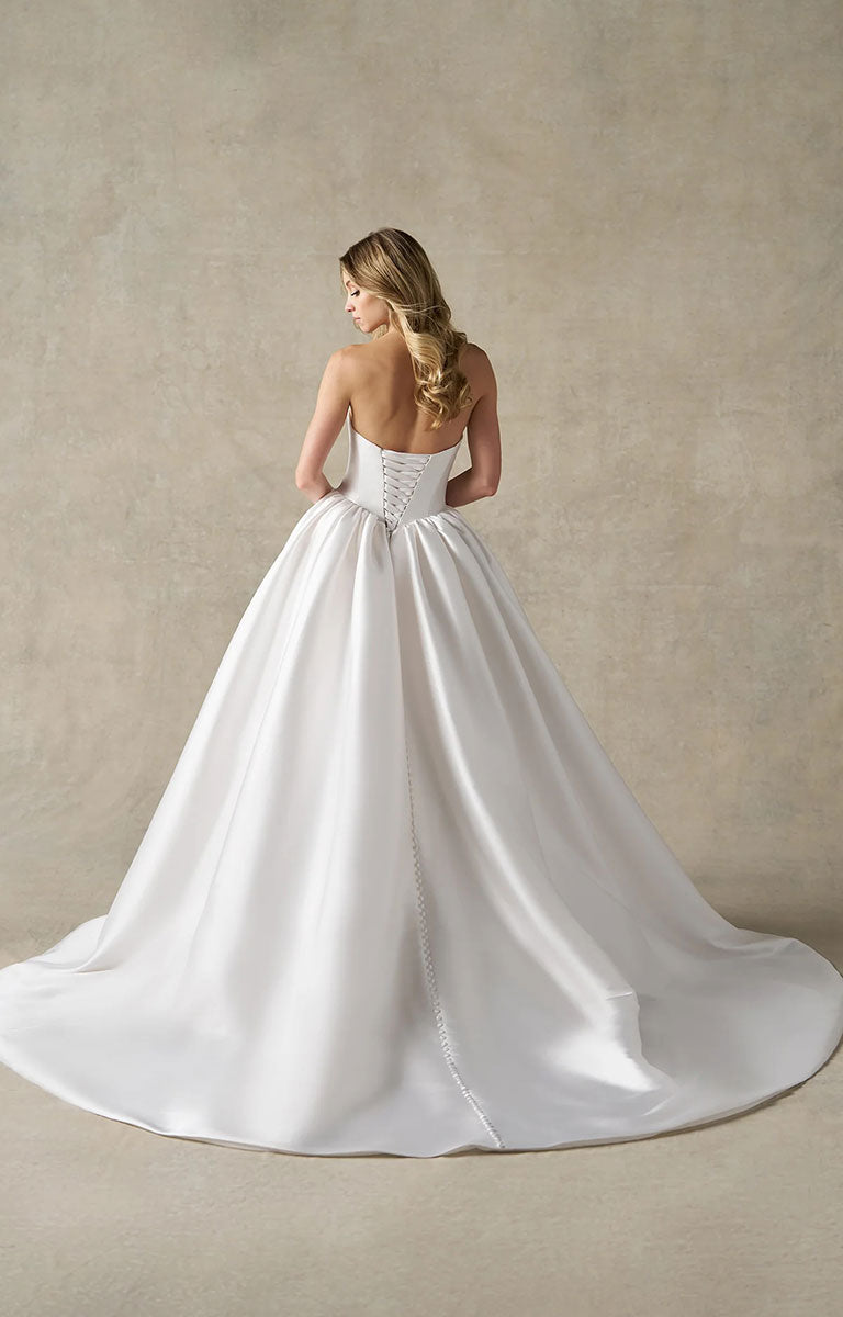 A woman modeling A1492 by Allure Bridals - Mikado Ball Gown with Basque Waist, Sweetheart Neckline and Detachable Panniers from the back, showing a structured corset-style waist and a full satin train, against a warm beige textured studio backdrop.
