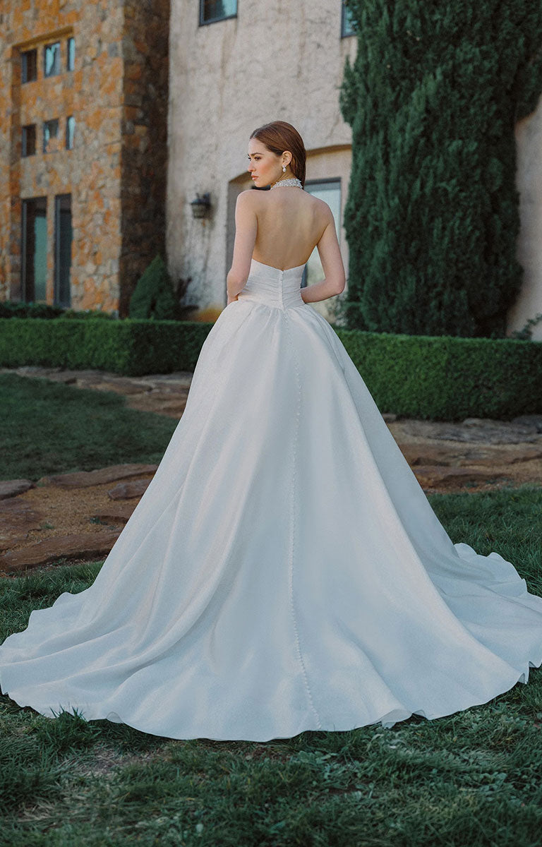 A woman wearing A1498 by Allure Bridals - Mikado Ball Gown with Beaded Halter Neckline and Pearl Detailing stands outdoors on grass in front of a stone building and greenery, photographed from the back to show the open back and full train of the dress.