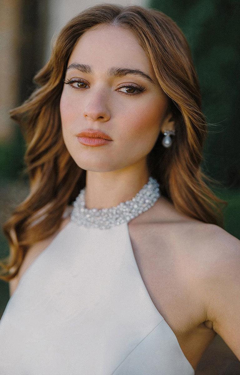 A woman with wavy brown hair A1498 by Allure Bridals - Mikado Ball Gown with Beaded Halter Neckline and Pearl Detailing and a sparkling pearl choker necklace, photographed in warm outdoor light with a blurred green background.