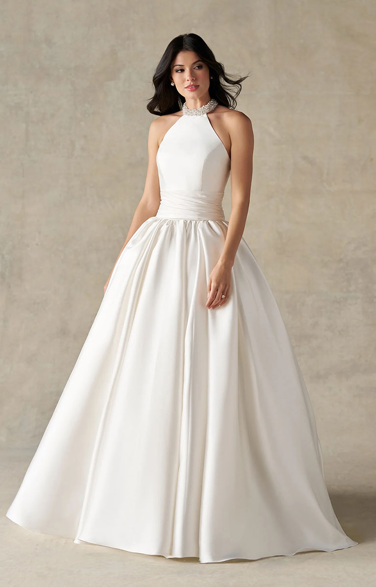 A woman modeling A1498 by Allure Bridals - Mikado Ball Gown with Beaded Halter Neckline and Pearl Detailing with a structured bodice and a full, flowing skirt in an ivory or off-white satin-like fabric, set against a textured beige studio backdrop.