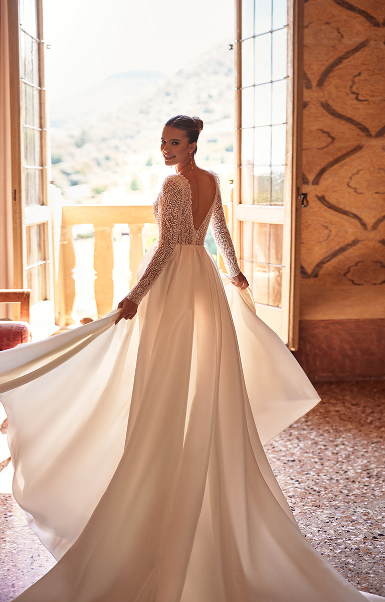Woman in a white wedding dress with long sleeves standing in a room with large windows.