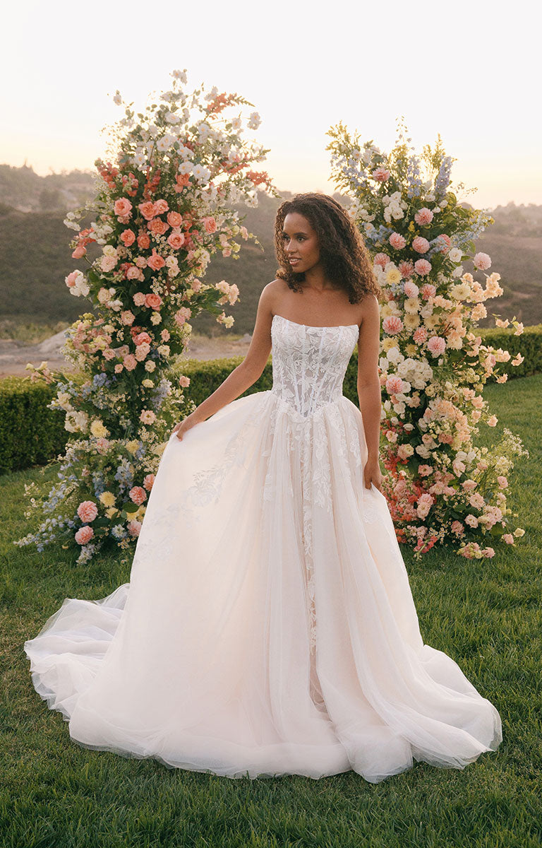 Woman in A1408 by Allure Bridal - Strapless Sequin Lace and Tulle Ball Gown Wedding Dress standing in front of floral arrangements outdoors.