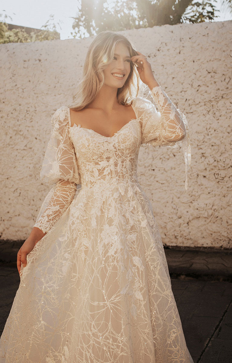 Woman wearing a lace wedding dress with long sleeves against a textured wall.