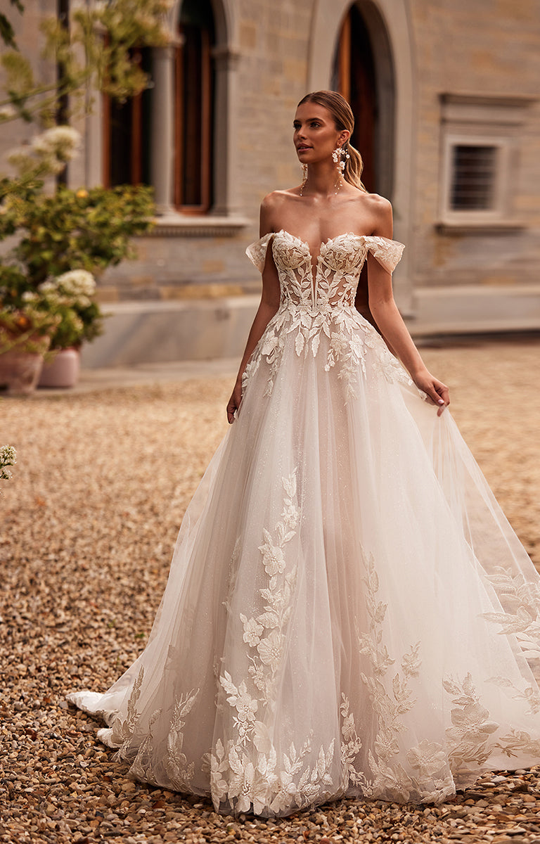 Woman in a white wedding dress with floral details standing in front of a stone building.