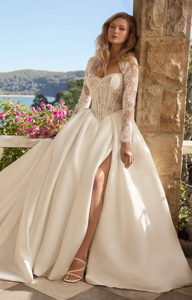 A woman models Bree Y3221 by Sophia Tolli - Corset Ball Gown Wedding Dress with Satin Skirt and Detachable Lace Jacket with a lace bodice, long lace sleeves, and a high thigh slit, standing outdoors beside a stone pillar with a garden and water in the background.