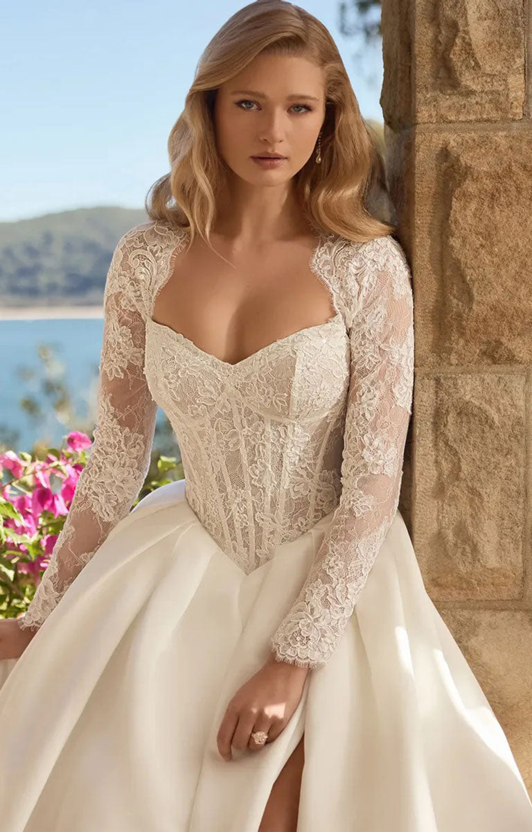 A woman wearing Bree Y3221 by Sophia Tolli - Corset Ball Gown Wedding Dress with Satin Skirt and Detachable Lace Jacket with a sweetheart neckline and full skirt, posed outdoors against a stone wall with water and flowers in the background.
