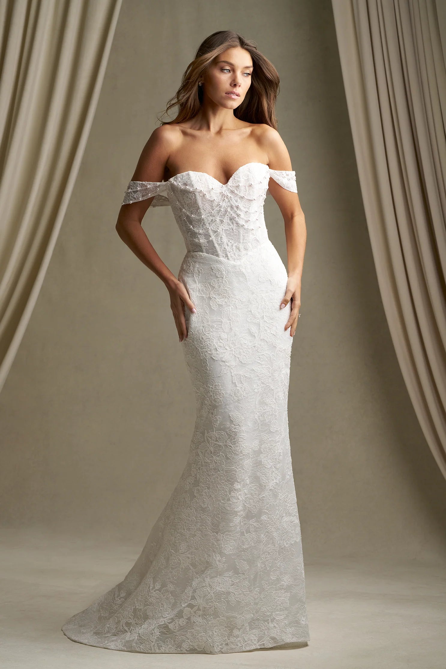 Woman wearing C803 by Allure Couture - Off-the-Shoulder Embroidered Lace Sheath Wedding Dress with Pearls against a beige curtain backdrop