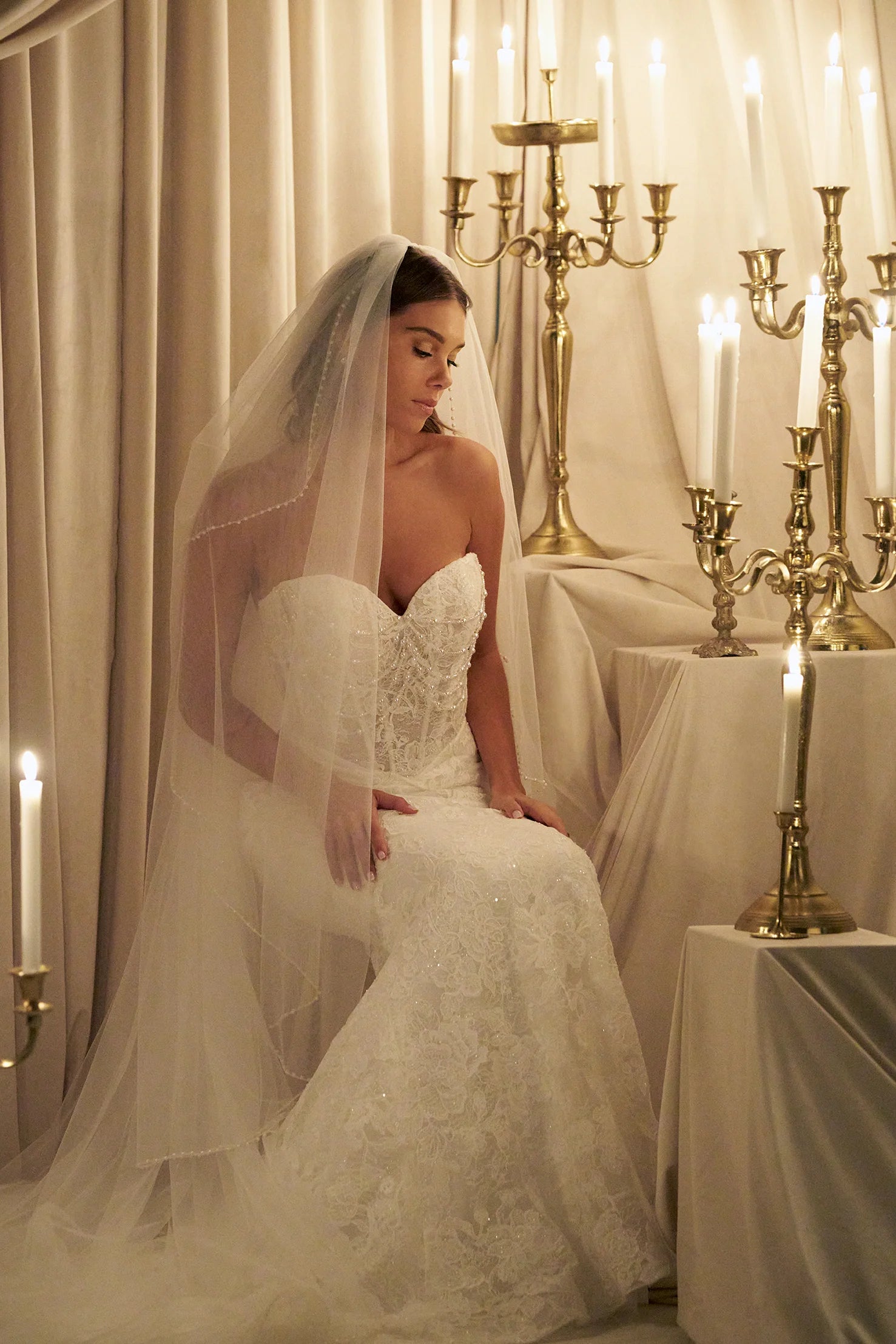Woman in C803 by Allure Couture - Off-the-Shoulder Embroidered Lace Sheath Wedding Dress with Pearls sitting under a draped arch with candles and candelabras.