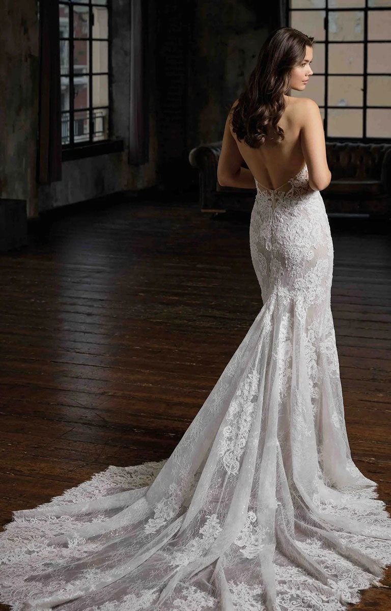 A woman wearing Pascale CT748 by Eddy K Milano Couture - Lace Fit-and-Flare Wedding Dress with Halter Neckline with a long train, photographed from the back in a dimly lit indoor room with dark wood floors and windows.