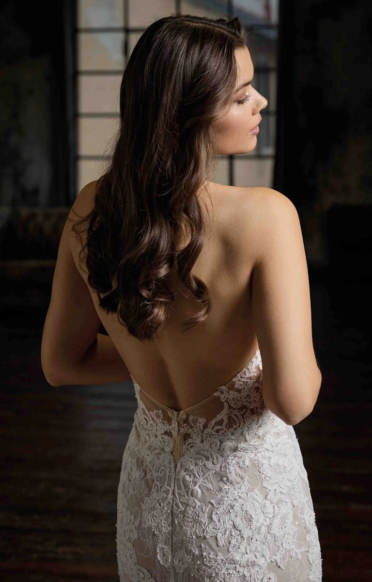 A woman with long, wavy brown hair is shown from behind wearing Pascale CT748 by Eddy K Milano Couture - Lace Fit-and-Flare Wedding Dress with Halter Neckline with a low open back, set against a dark, moody indoor background.