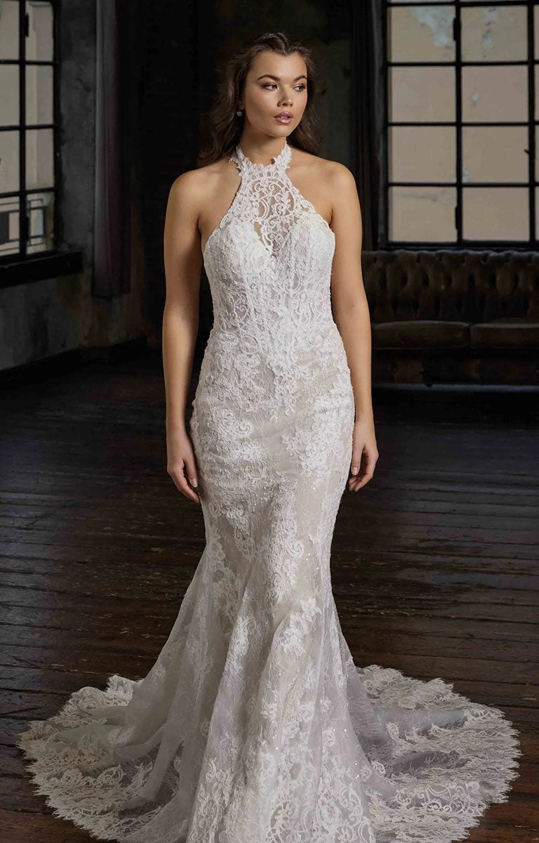 A woman modeling Pascale CT748 by Eddy K Milano Couture - Lace Fit-and-Flare Wedding Dress with Halter Neckline with a high halter neckline and a long sheer train, photographed in a dimly lit indoor room with dark wood floors and windows.
