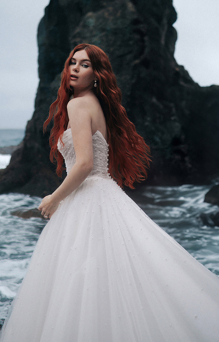 Woman with red hair wearing the D440 Ariel - Disney Fairytale Weddings Wedding Dress standing by the ocean.