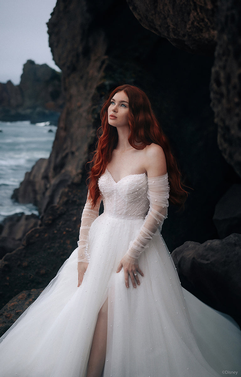 Woman wearing the D440 Ariel - Disney Fairytale Weddings Wedding Dress with optional long sleeves standing on a rocky coastal landscape.