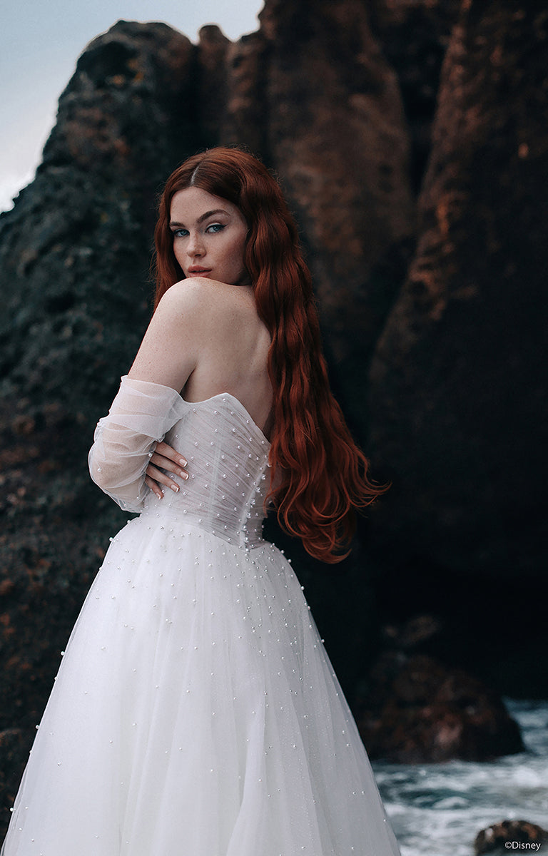 Woman with red hair wearing the D440 Ariel - Disney Fairytale Weddings Wedding Dress in front of a rocky coastal background.