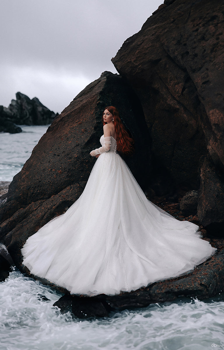 Woman with red hair wearing the D440 Ariel - Disney Fairytale Weddings Wedding Dress standing on rocks in front of the ocean.
