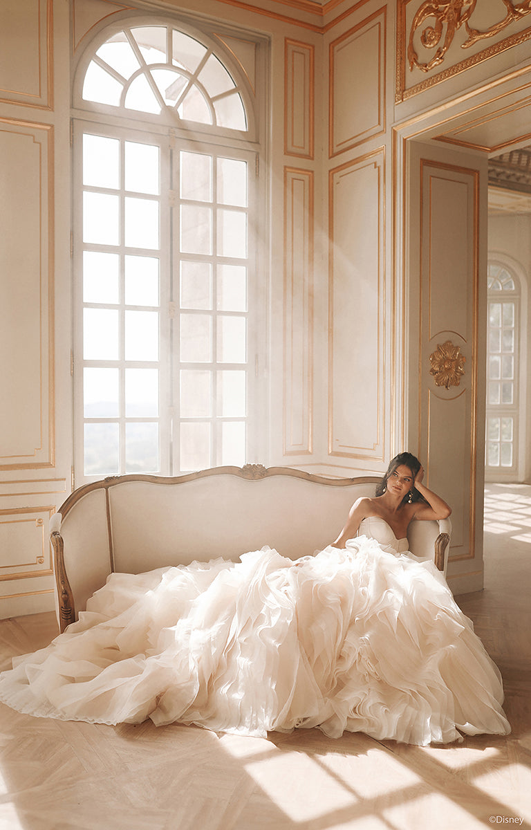 Woman in DP422 Belle - Disney Fairytale Weddings Wedding Dress sitting on a couch in a sunlit room with ornate decor.