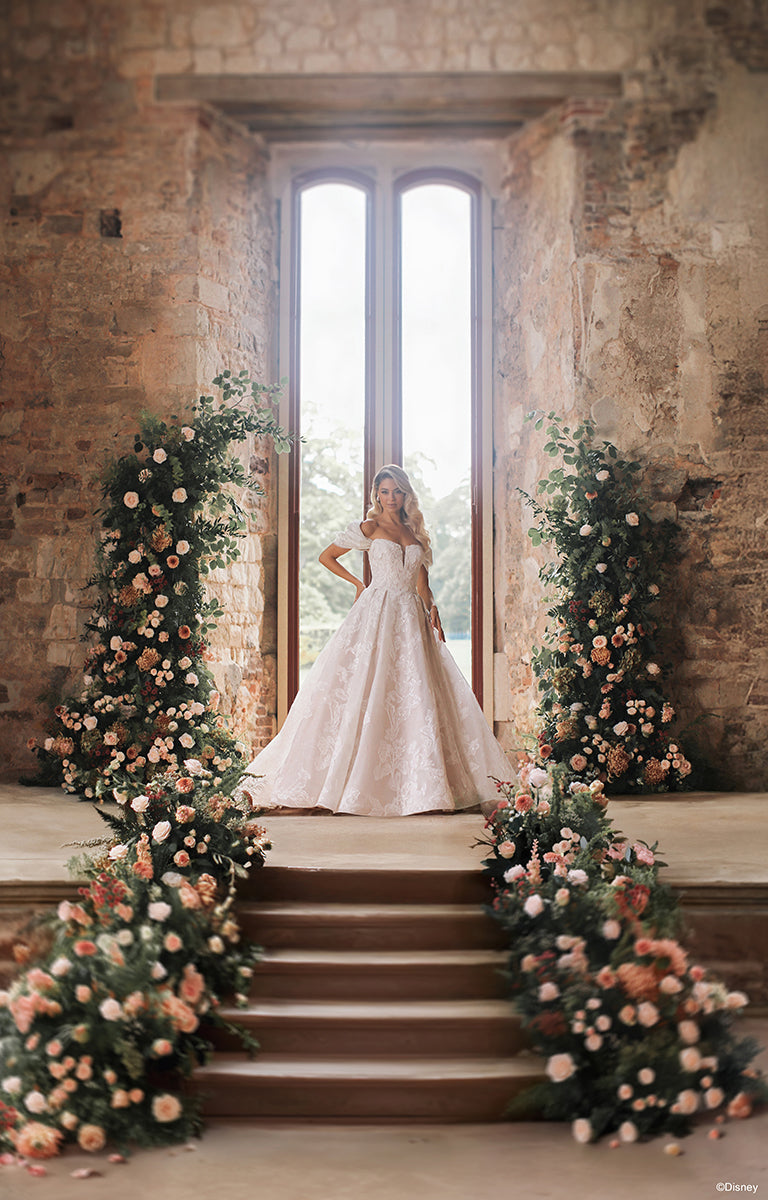 Bride in DP451 Sleeping Beauty - Disney Fairytale Weddings Wedding Dress standing on a stone staircase with floral decorations in a rustic setting.