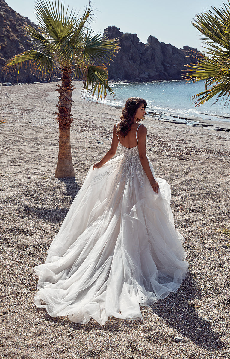 Woman in a white wedding dress standing on a sandy beach with palm trees and water in the background.