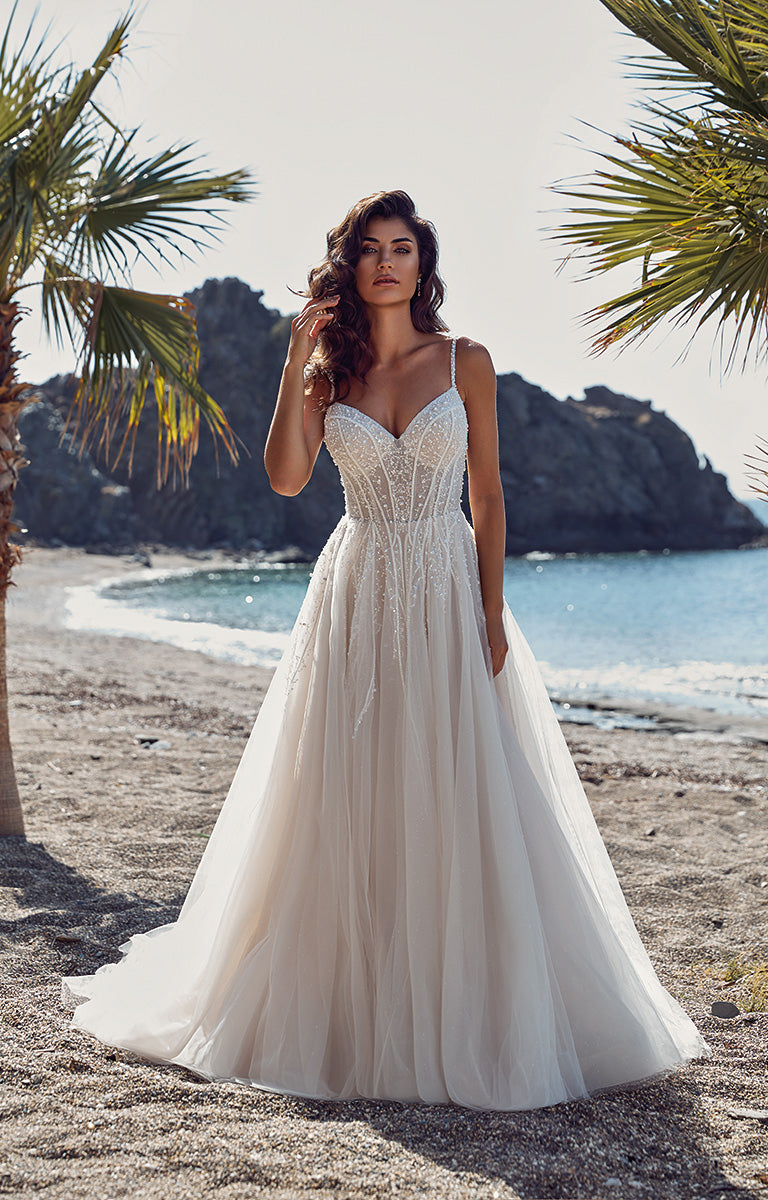 Woman in a white wedding dress standing on a beach with palm trees and rocky cliffs in the background.