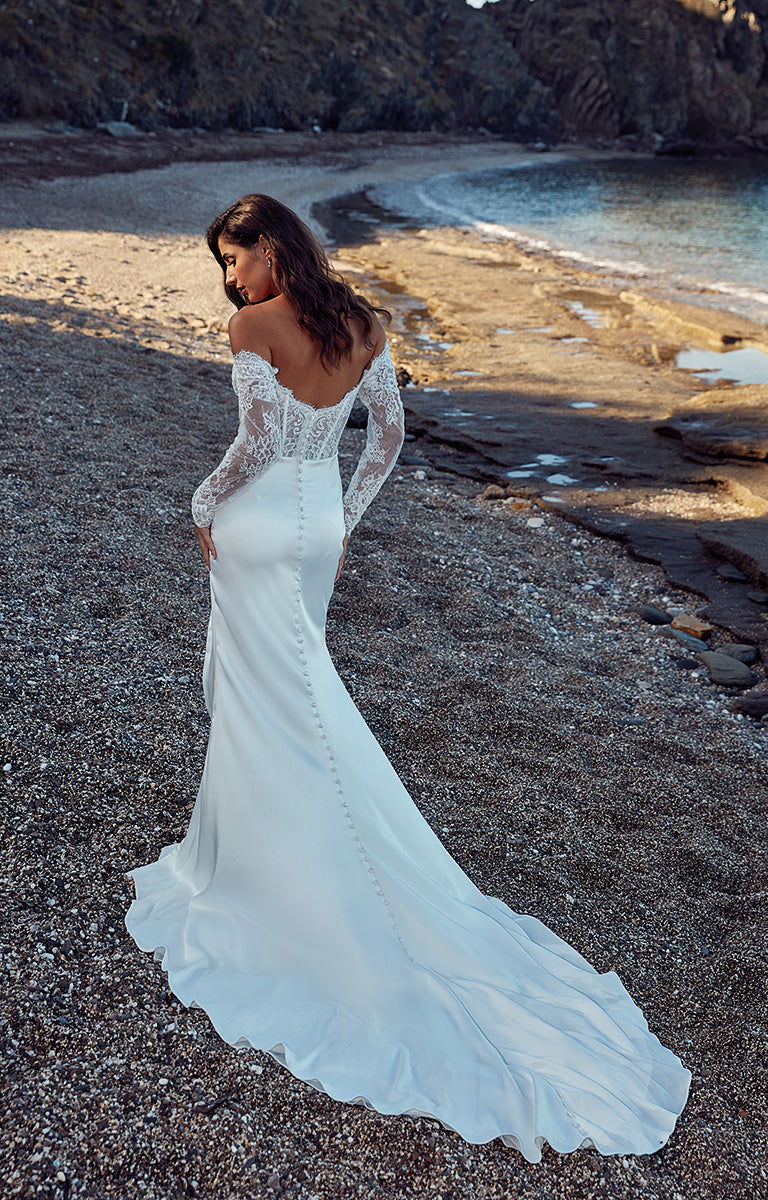 Woman in a white wedding dress standing on a pebbly beach with water and rocks in the background.
