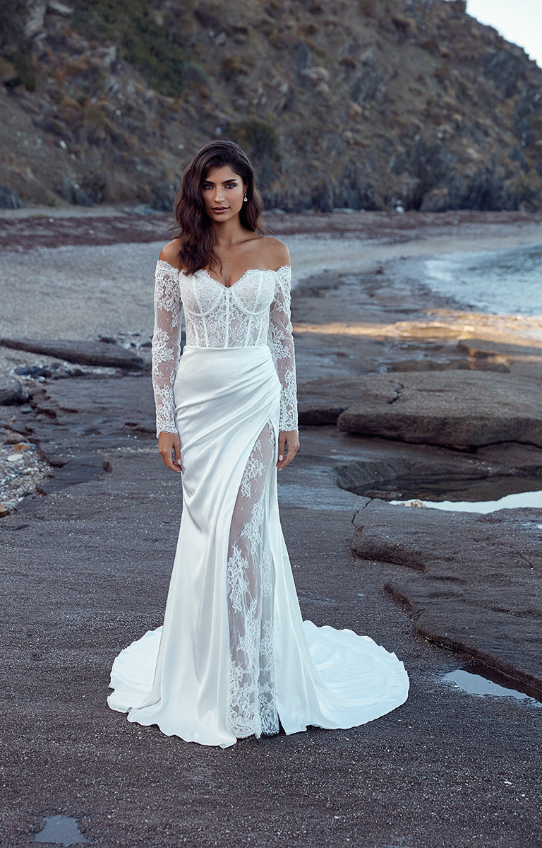 Woman in a white lace wedding dress standing on a rocky beach.