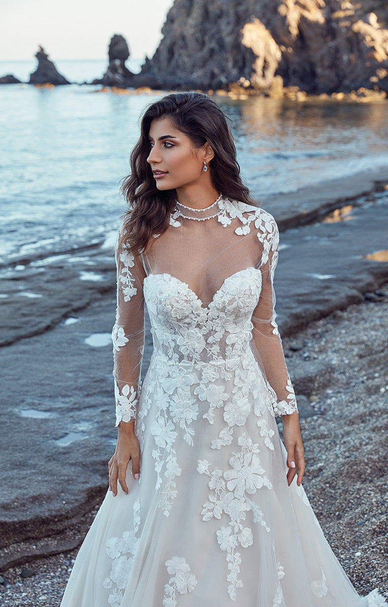 Woman in a white lace wedding dress standing on a beach with rocky formations in the background.
