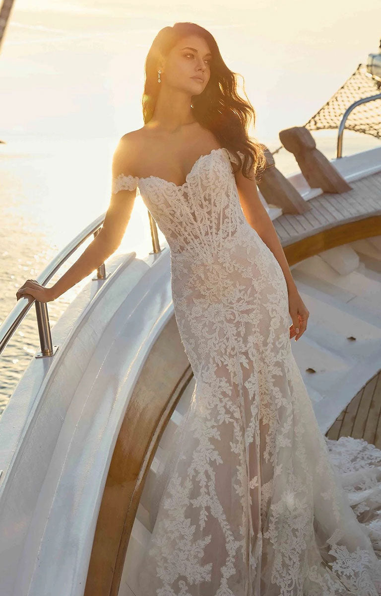 A woman wearing Rio DR2706 by Eddy K Dreams - Ivory and Lavender Lace Fit-and-Flare Wedding Dress stands on the deck of a yacht at sunset, with water and warm sunlight in the background.