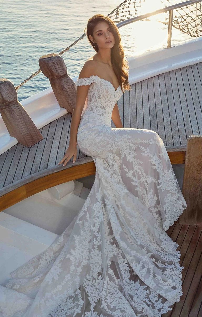 A woman wearing Rio DR2706 by Eddy K Dreams - Ivory and Lavender Lace Fit-and-Flare Wedding Dress sits on the wooden deck of a yacht with the ocean in the background.