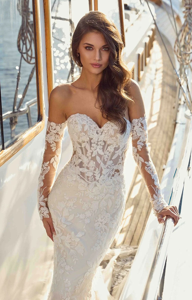 A woman wearing Neha DR2710 by Eddy K Dreams - Beaded Lace and Sequin Fit-and-Flare Wedding Dress stands on a sunlit boat deck, with the ship's rigging and water visible in the background.