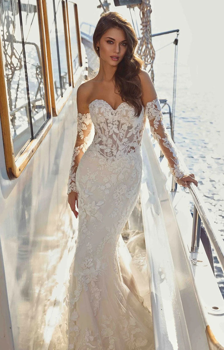 A woman wearing Neha DR2710 by Eddy K Dreams - Beaded Lace and Sequin Fit-and-Flare Wedding Dress stands on the deck of a boat with water and bright sunlight in the background.