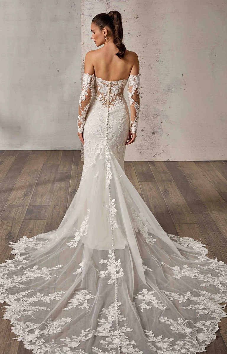A woman modeling Neha DR2710 by Eddy K Dreams - Beaded Lace and Sequin Fit-and-Flare Wedding Dress from the back, showing an off-the-shoulder lace design, fitted silhouette, and long sheer lace sleeves with a dramatic train on a rustic indoor set.