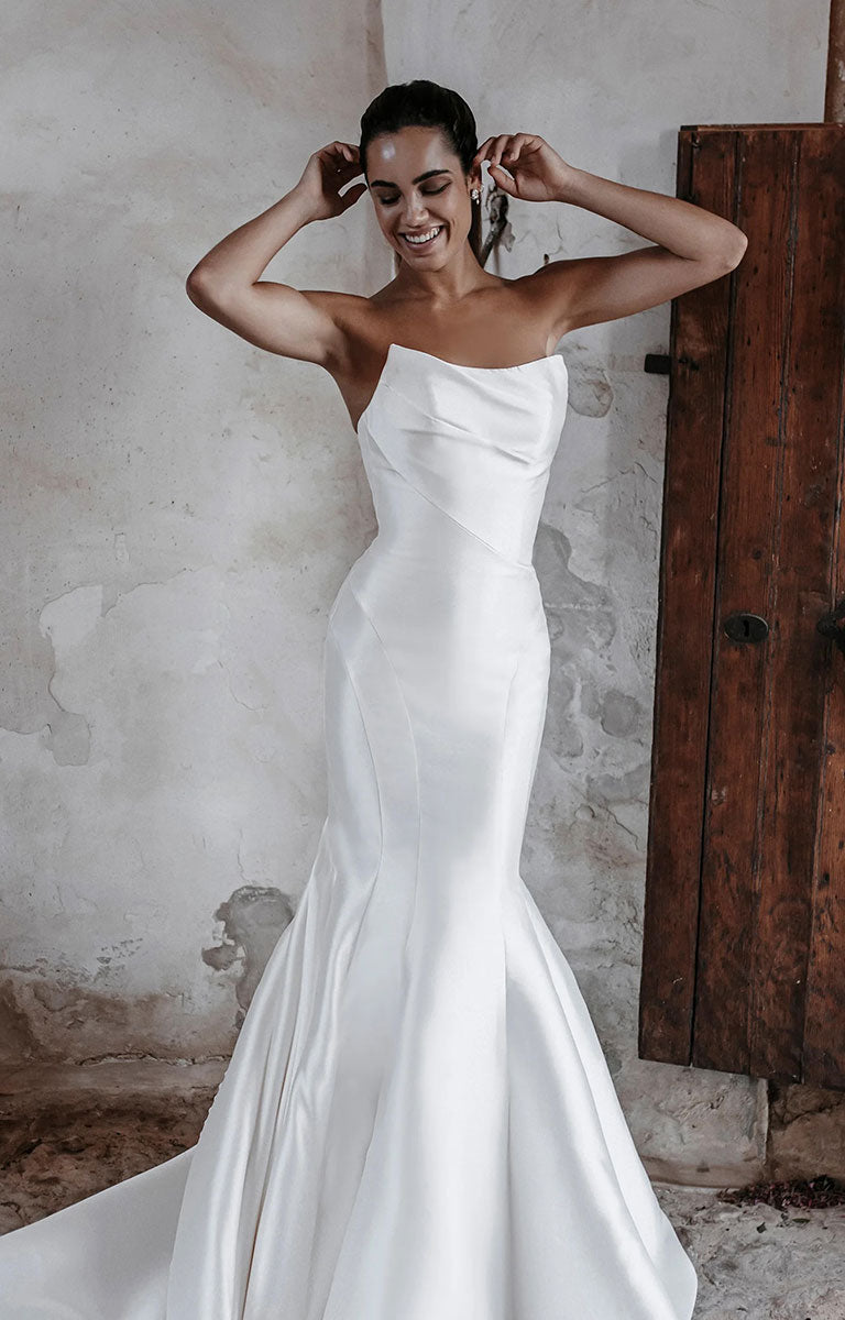 Woman in Carmen E215 by Abella Bridal - Strapless Minimalist Wedding Dress with Dramatic Neckline standing against a rustic wall.