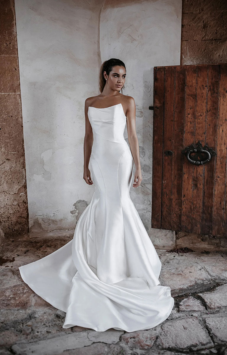 Woman in Carmen E215 by Abella Bridal - Strapless Minimalist Wedding Dress with Dramatic Neckline standing in front of a rustic wooden door.