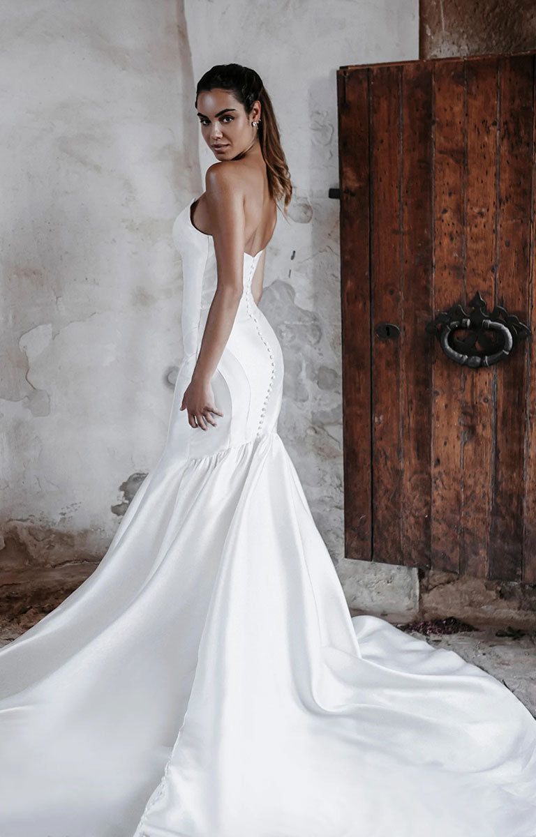 Woman in Carmen E215 by Abella Bridal - Strapless Minimalist Wedding Dress with Dramatic Neckline standing in front of a rustic wooden door.