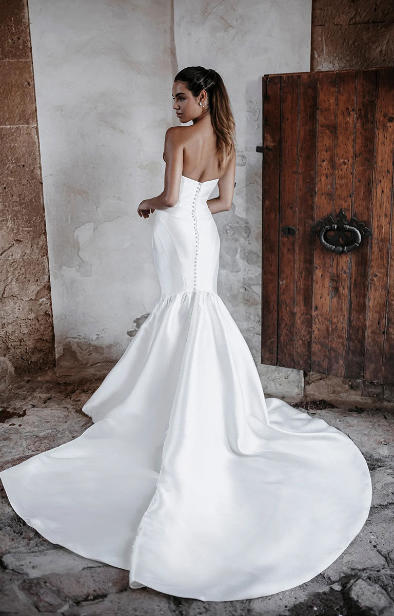 Woman in Woman in Carmen E215 by Abella Bridal - Strapless Minimalist Wedding Dress with Dramatic Neckline standing in front of a rustic wooden door. 