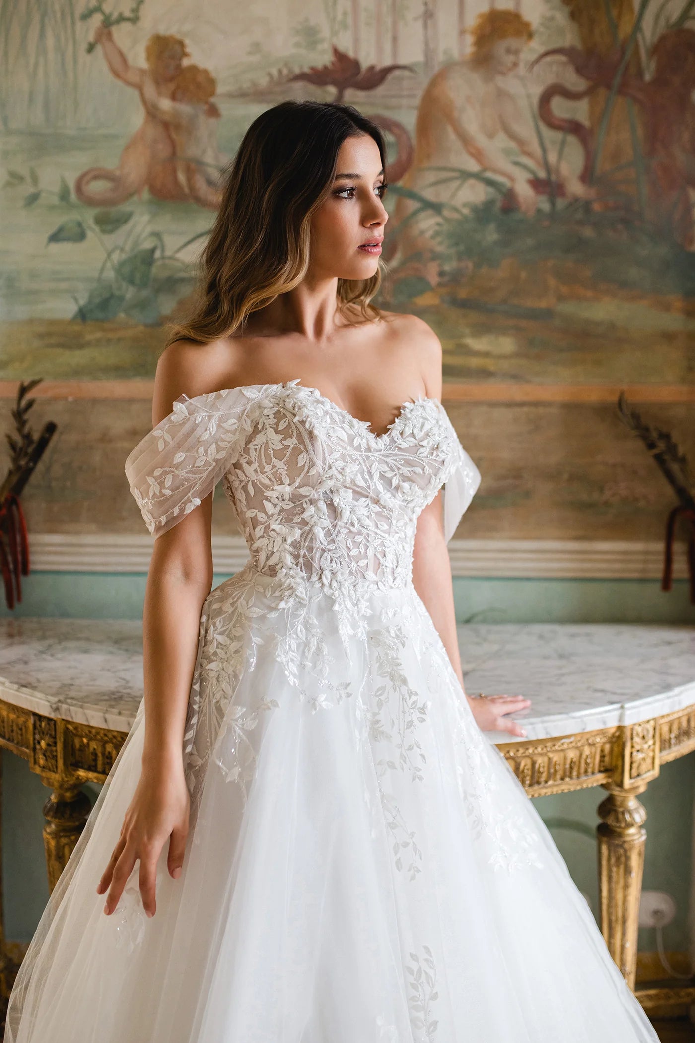Woman in a white lace wedding dress standing in an ornate room with classical frescoes.