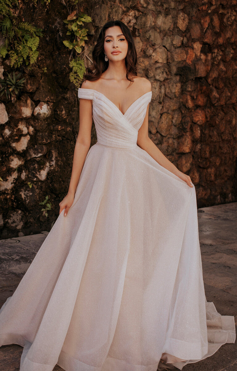 Woman in a white off-shoulder wedding dress standing against a stone wall.
