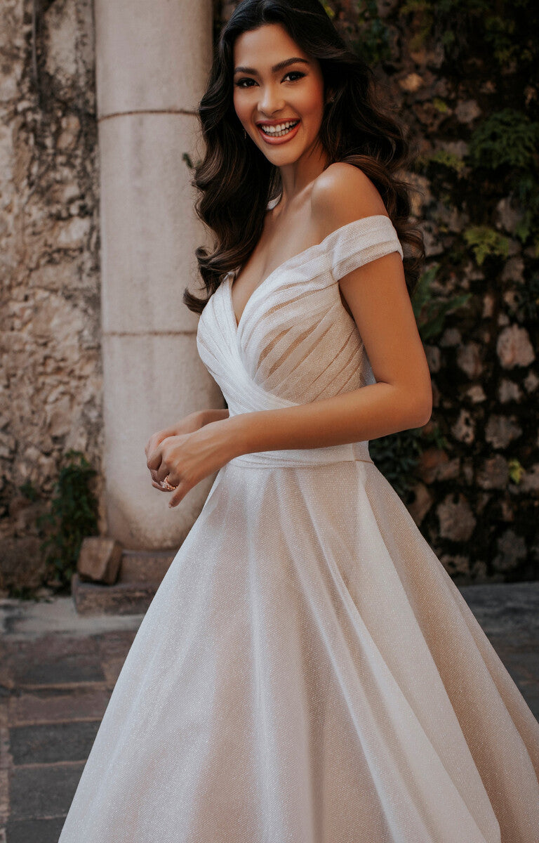 Woman in a white off-shoulder dress standing outdoors with a stone wall and plants in the background.