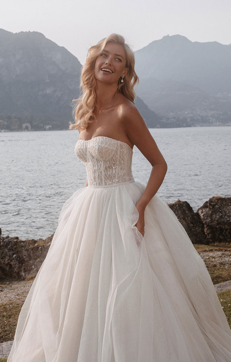 Woman wearing Stella E405 by Abella - Strapless Sparkle Tulle Ballgown Wedding Dress with a lake and mountain backdrop.