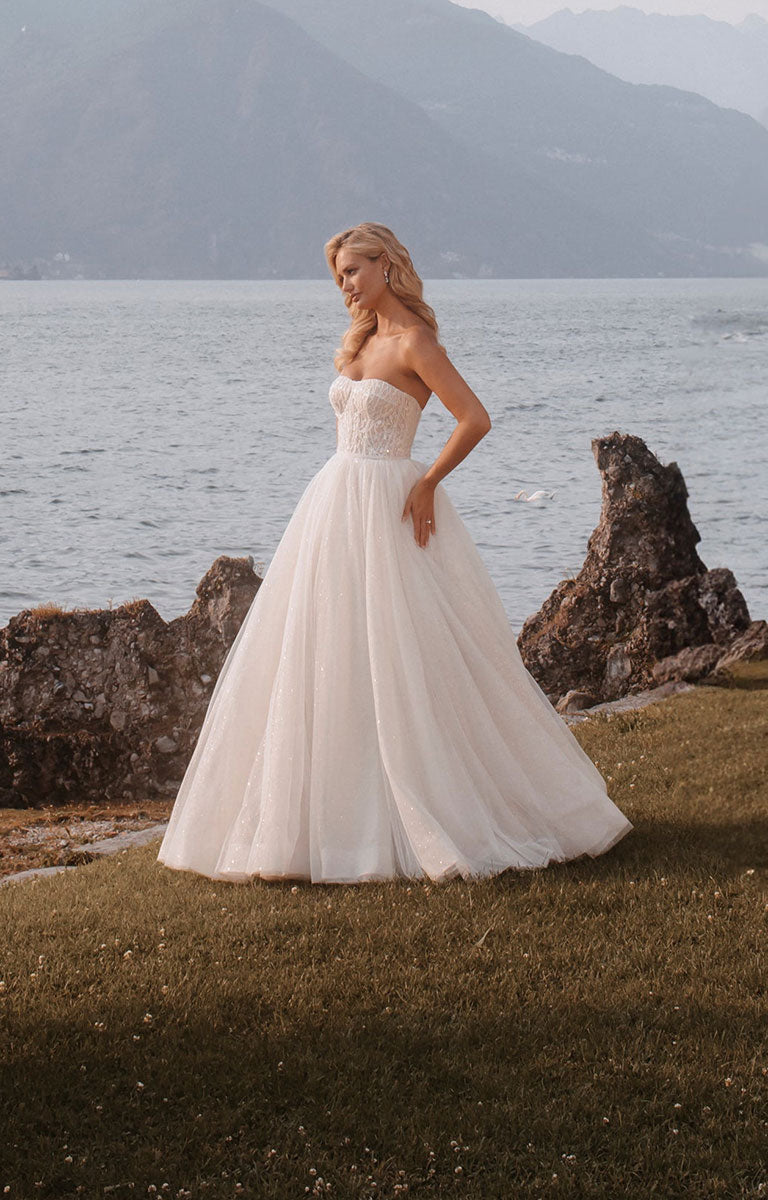 Woman wearing Stella E405 by Abella - Strapless Sparkle Tulle Ballgown Wedding Dress with a lake and mountain backdrop.
