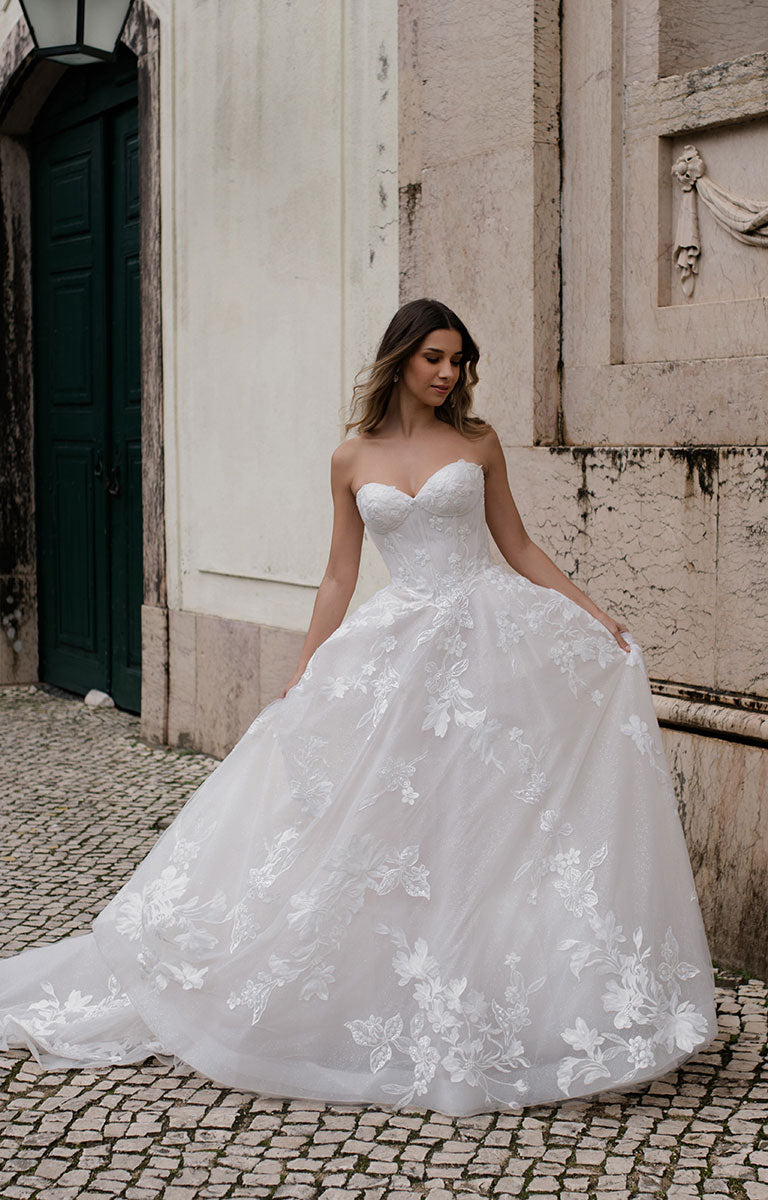 Woman in a white wedding dress with floral details standing in front of a stone building.