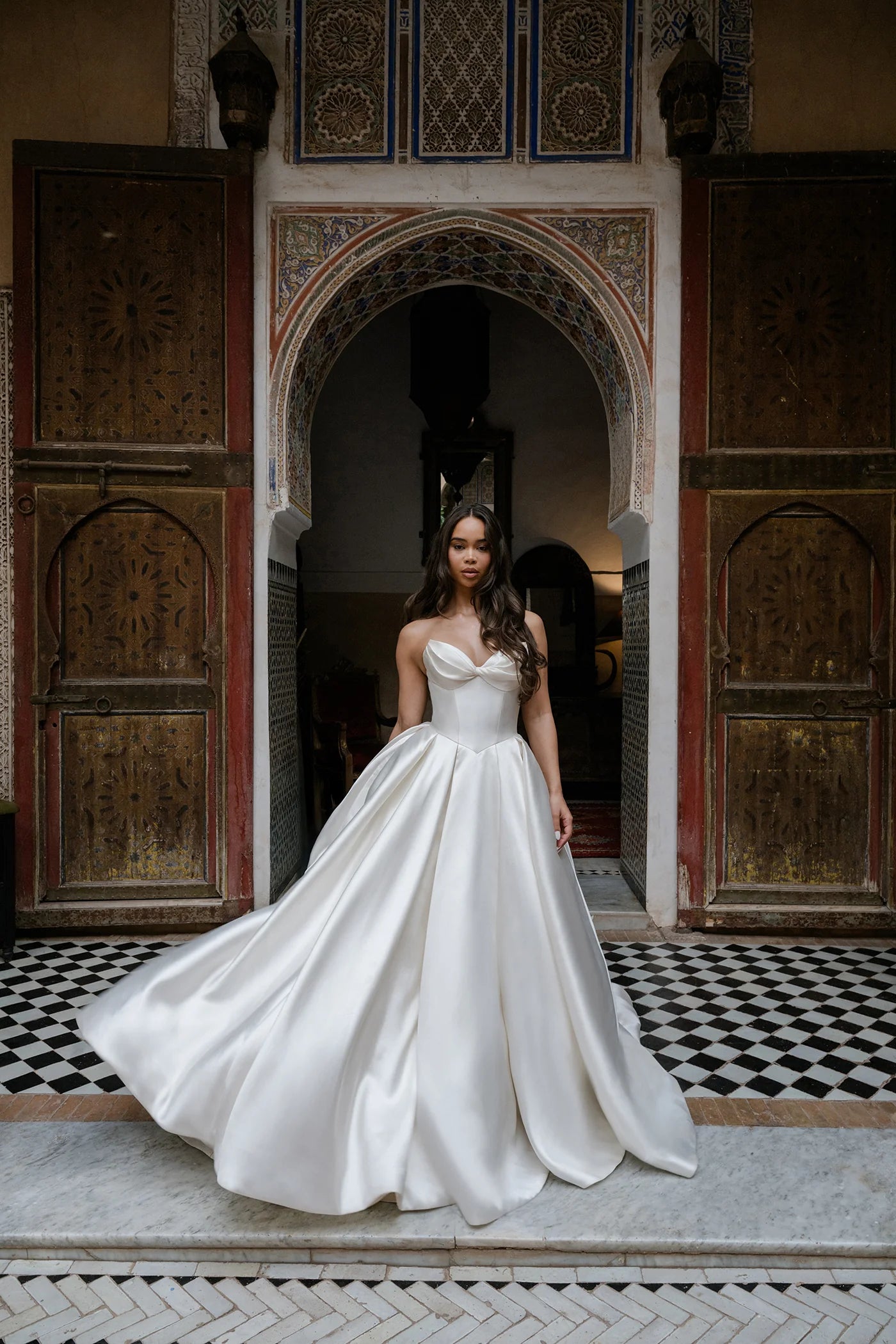 Woman in a white wedding dress standing in an ornate room with wooden doors and intricate patterns.