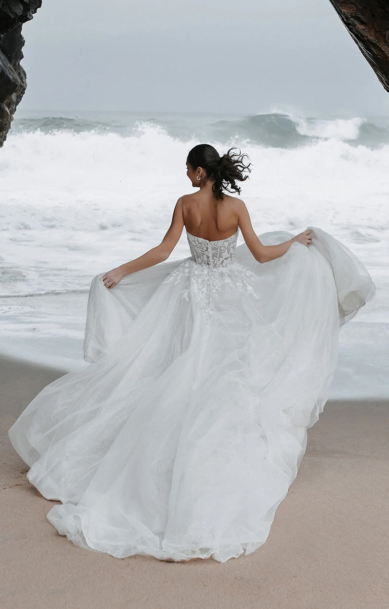 Woman in Benedita E461 by Abella Bridal - Beaded Lace A-Line Tulle Wedding Dress standing on a beach with waves in the background