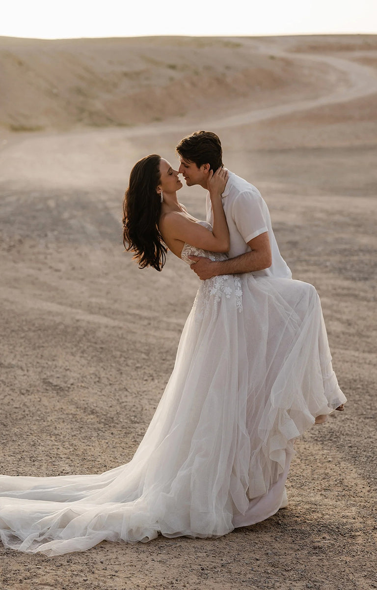 Couple in wedding attire embracing in a desert setting. The woman is wearing Benedita E461 by Abella Bridal - Beaded Lace A-Line Tulle Wedding Dress.